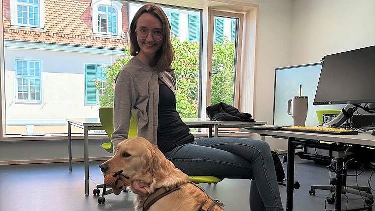 Eine Frau sitzt auf einem Stuhl im Klassenzimmer, vor ihr sitzt ein hellbrauner Hund mit Plüschtier im Maul. Hinter ihr ist ein großes Fenster mit Blick auf das gegenüberliegende rosafarbene Backsteingebäude.
