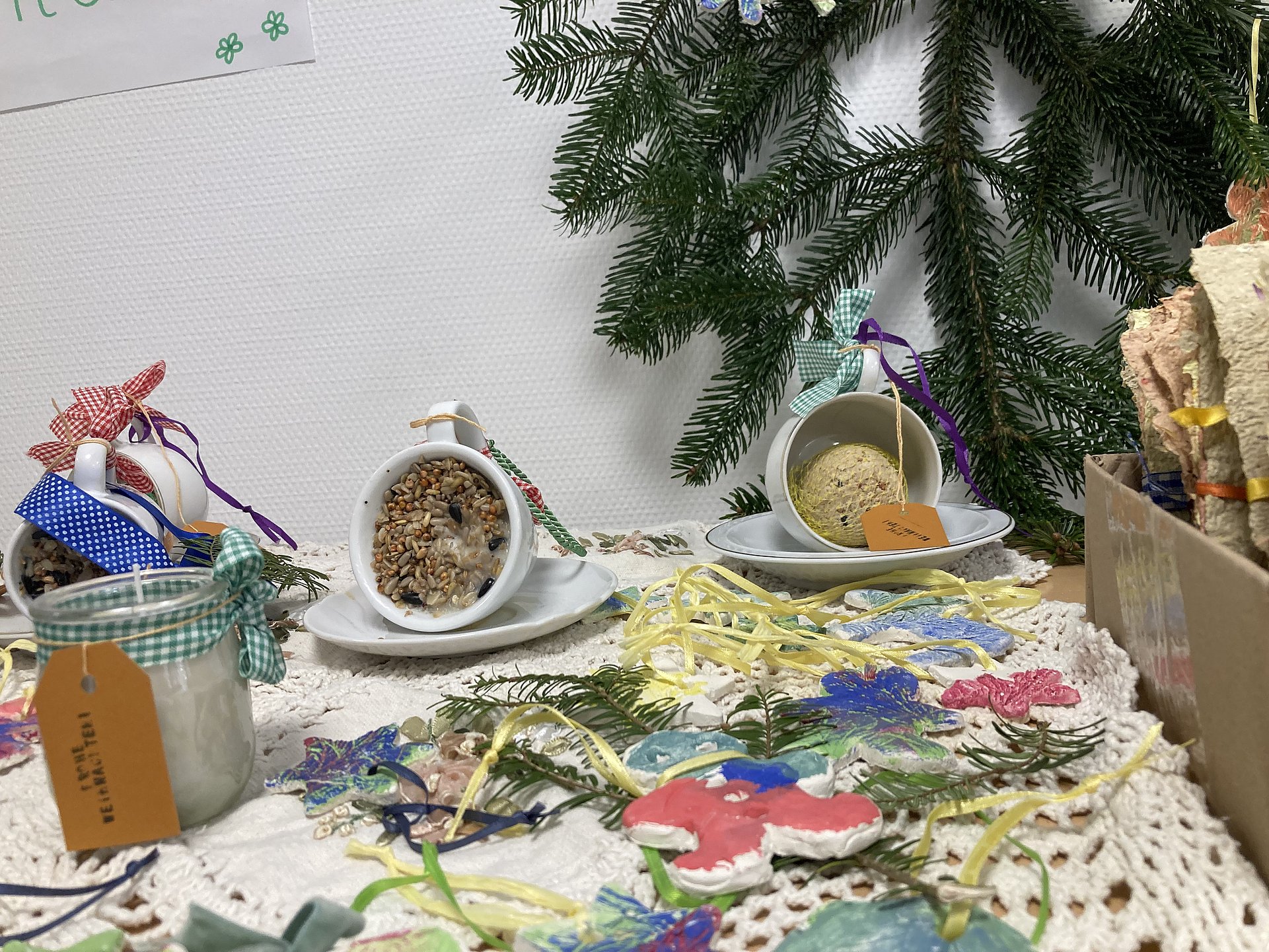 Auf einem Tisch liegen Meisenknödel in Keramiktassen, Bunte Anhänger und eine Duftkerze in einem Glas