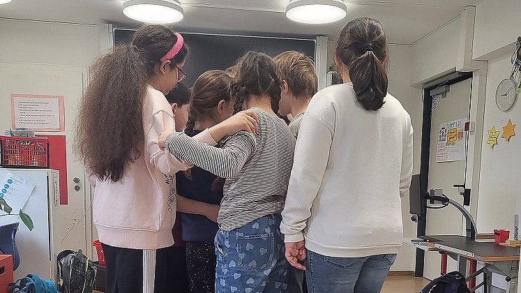 Mehrere Kinder stehen gemeinsam auf einer kleinen Holzbrücke im Klassenzimmer. Sie halten sich aneinander fest mit dem Rücken zur Kamera.
