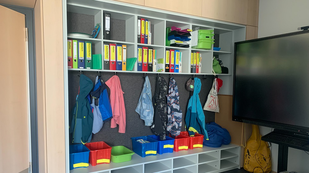 In einem Klassenzimmer ist die Garderobe gefüllt mit Ordnern, Kisten und Jacken, rechts daneben steht ein schwarzes Smartboard.