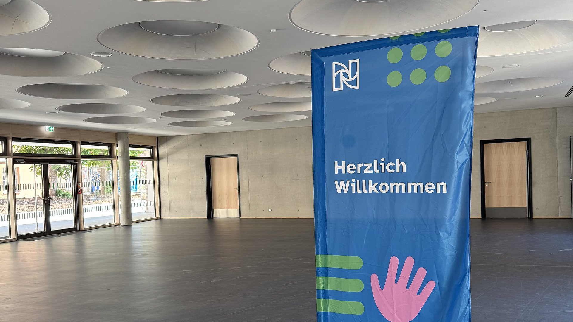 In einem großen Raum mit Fensterfront und großen Lichtern an der Decke steht eine blaue Fahne mit "Herzlich willkommen" darauf.