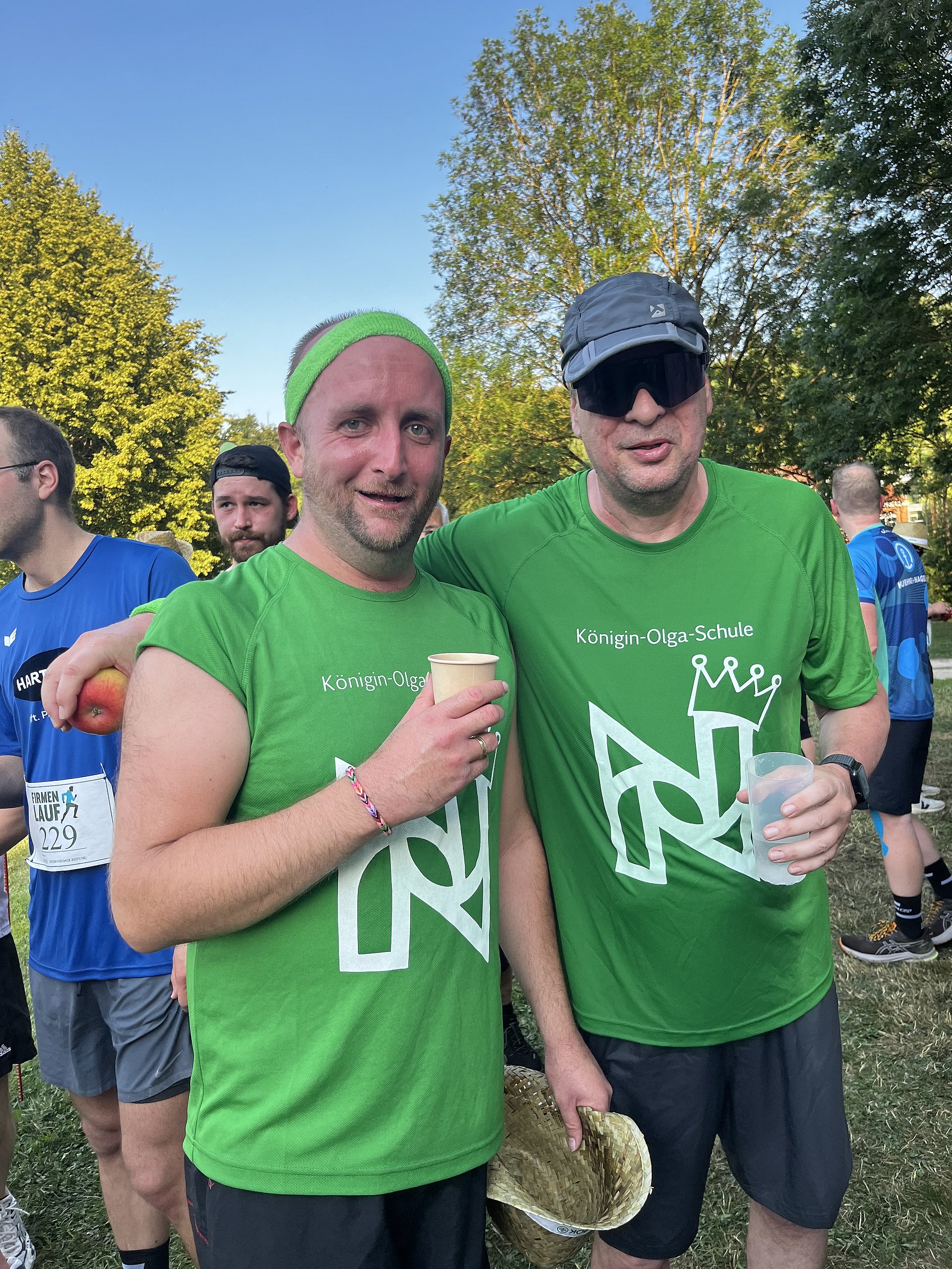 Zwei Männer in leuchtend grünen Laufshirts nach einem Laufveranstaltung. Beide stehen dicht nebeneinander im Freien auf einer Wiese, im Hintergrund sind weitere Läufer, Bäume und Abendlicht.