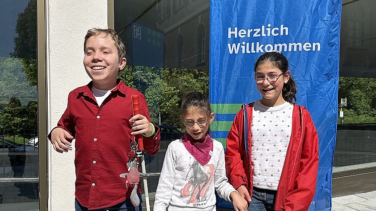 Ein Junge mit Blindenstock und zwei Mädchen stehen vor dem Gebäude nebeneinander, sie lachen und schauen in die Kamera. Hinter ihnen steht eine blaue Fahne mit "Herzlich willkommen".