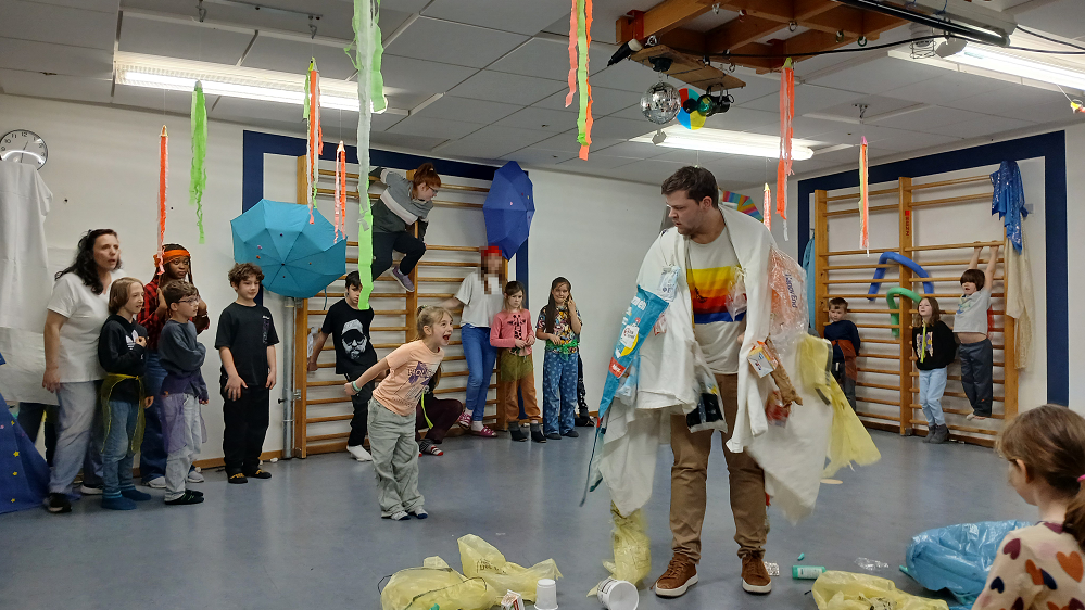 Ein Mann steht in der Mitte eines Raums, er trägt eine Art Umhang aus Müll, um ihn herum liegen Tüten und Plastikbecher. Um ihn herum stehen viele Kinder und Erwachsene und schauen ihn an. Ein Mädchen steht dicht neben ihm und schreit ihn an.