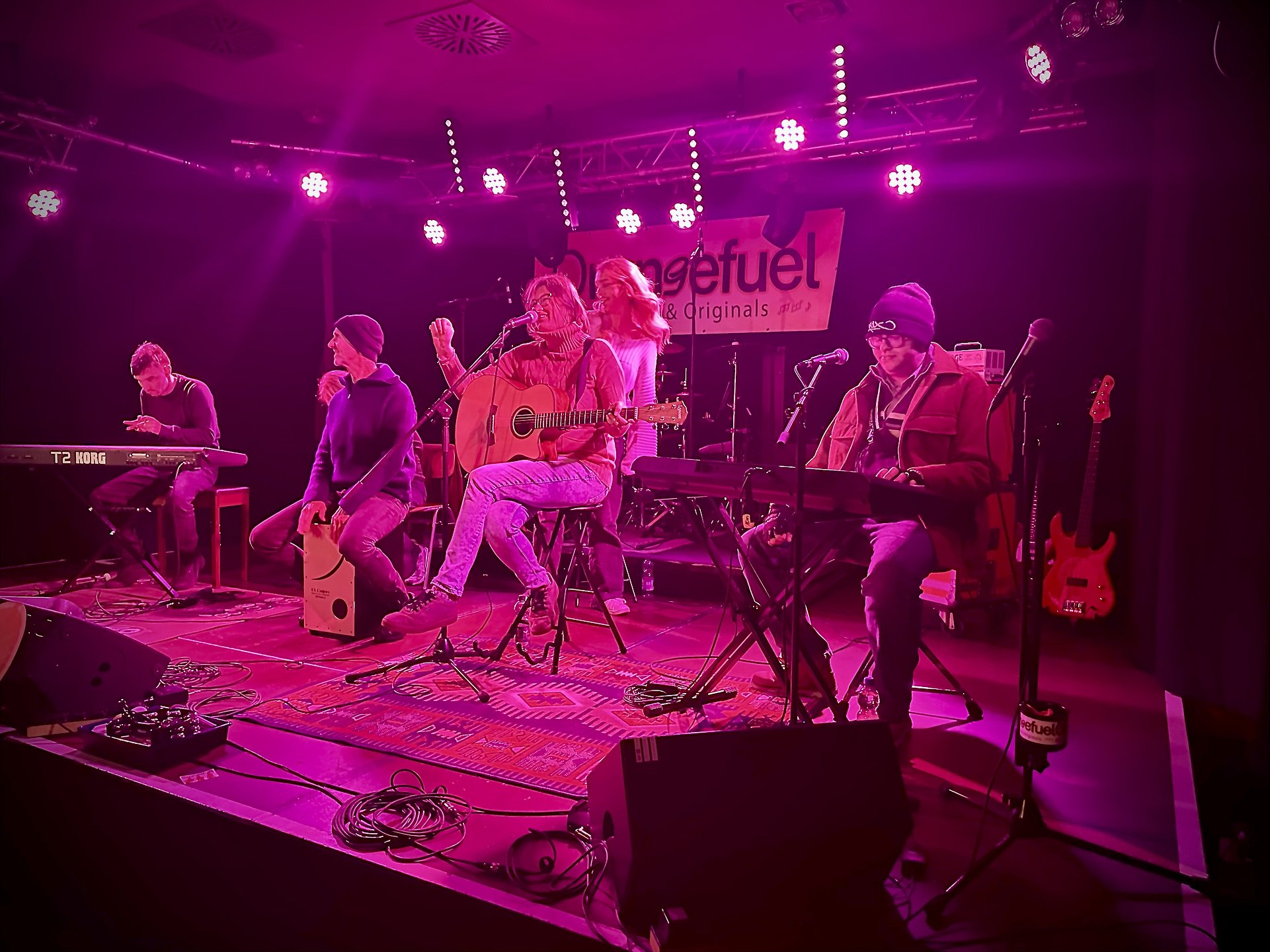 Eine Band steht in einem Pinken Licht Raum auf der Bühne und macht Musik