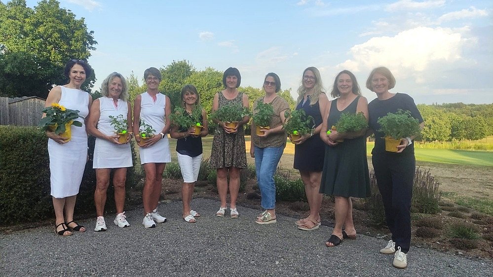 Ein Foto mit neun Frauen nebeneinanderstehend vor einer Wiese mit Bäumen. Sie tragen weiße und dunkle, schicke Kleidung und halten kleine Blumengestecke in der Hand. Sie lächeln alle in die Kamera. 
