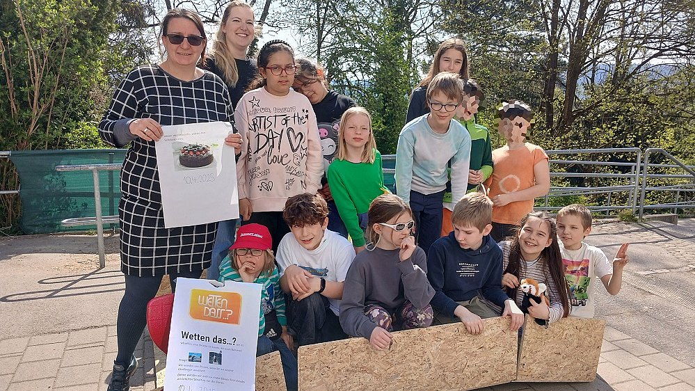 Mehrere Kinder und zwei Frauen stehen neben- und hintereinander auf dem Schulhof und lachen in die Kamera. Vor ihnen stehen Wellblechbretter aufgestellt.