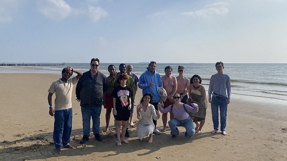 Eine Gruppe junger Erwachsener steht am Strand von Holland. Dahinter ist das Meer zu sehen mit einem blauen Himmel und kleinen Wolken