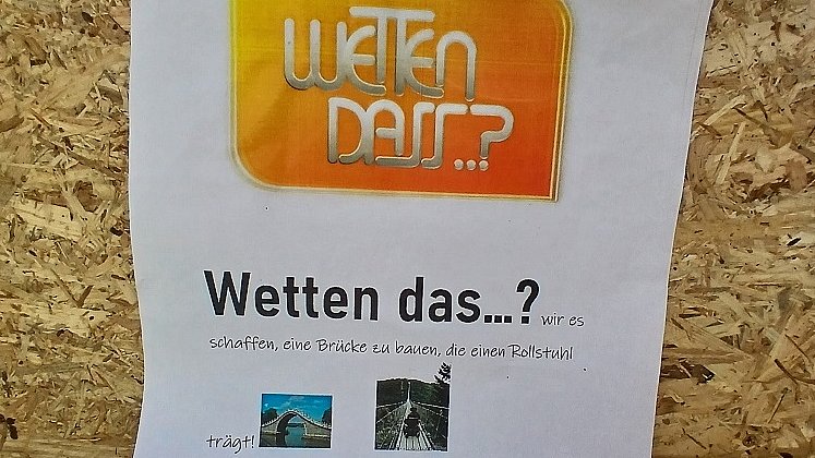 Ein Blatt Papier klebt auf einem Holzbrett. Auf dem Papier steht "Wetten dass...? wir es schaffen, eine Brücke zu bauen, die einen Rollstuhl trägt? Wir würden uns freuen, wenn ihr zu unserer Challenger annehmt. Gerne würden wir euch zu einem kleinen Fest einladen am letzten Schultag vor den Ferien.“