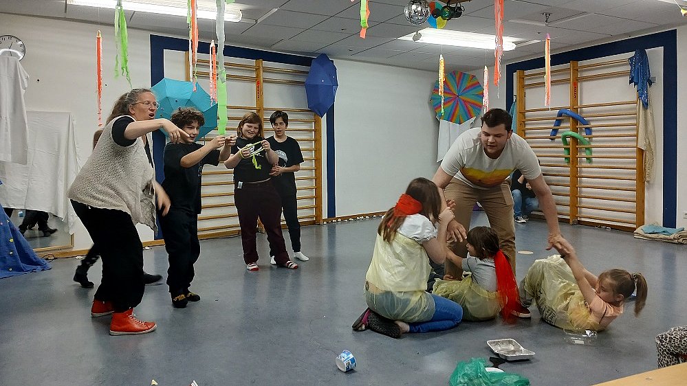 Ein Mann steht in der Mitte eines Raums um ihn herum sind zwei Kinder und eine junge Frau, sie tragen gelbe Müllsäcke um den Körper und zerren an dem Mann in der Mitte. Links daneben stehen vier Kinder und eine Frau in schwarzer Kleidung und schauen auf die Szene.