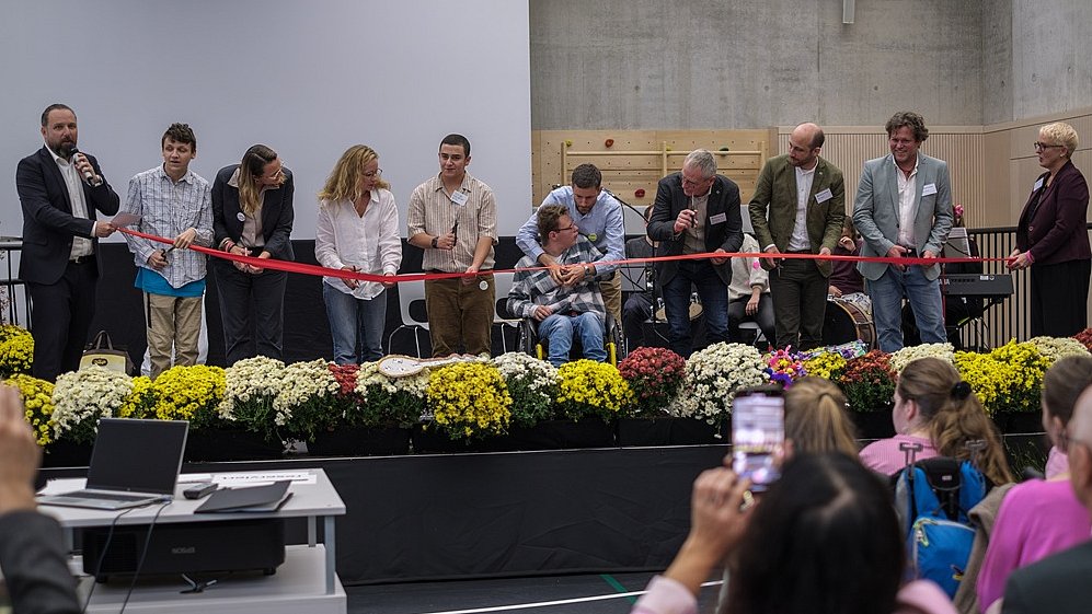 Auf der Bühne in der Sporthalle stehen 11 Personen nebeneinander, darunter Erwachsene und Jugendliche, einige davon im Rollstuhl. Sie halten ein langes rotes Band in der Hand sowie jede Person eine Schere.