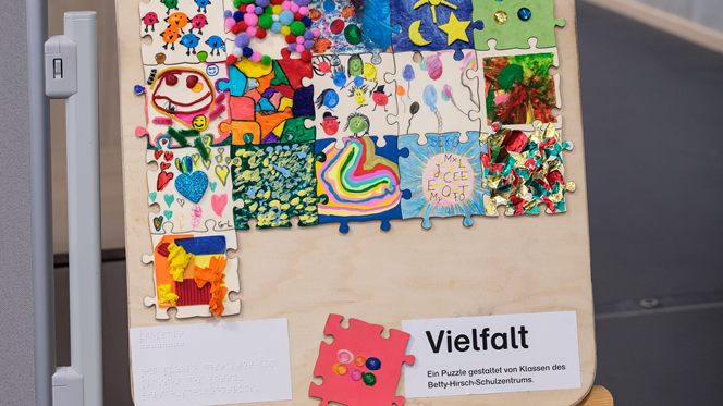 Auf einer Staffelei steht ein großes Bild, das mit Stoffen und Farben bunt gestaltet ist. Unten rechts in der Ecke steht "Vielfalt".