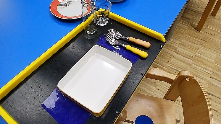 Auf einem blauen Tisch steht ein schwarzes Tablett mit gelbem Rahmen, darauf ein weißer Teller, Gelbes Besteck und zwei Gläser.
