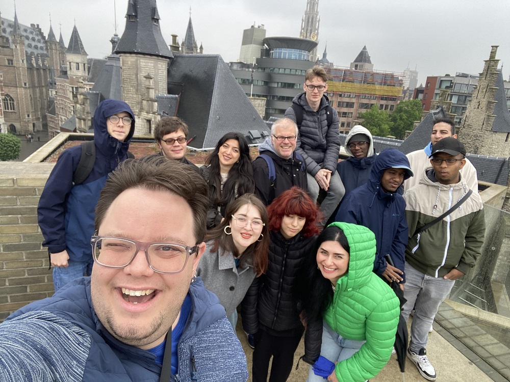 Eine Gruppe junger Erwachsener schaut lachend in die Kamera. Im Hintergrund sieht man die Stadt Antwerpen