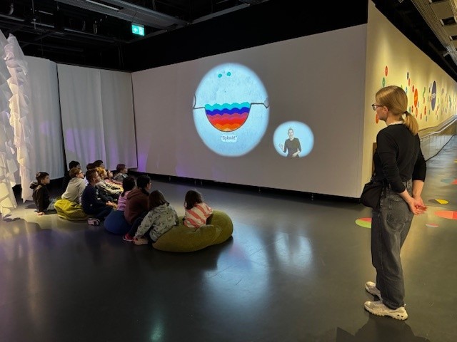 Die Schulklasse sitzt in der Ausstellung auf dem Boden auf Sitzkissen. Vor ihnen läuft ein Film auf einer großen Wand. Der Raum ist abgedunkelt. Rechts neben ihnen steht die Führerin