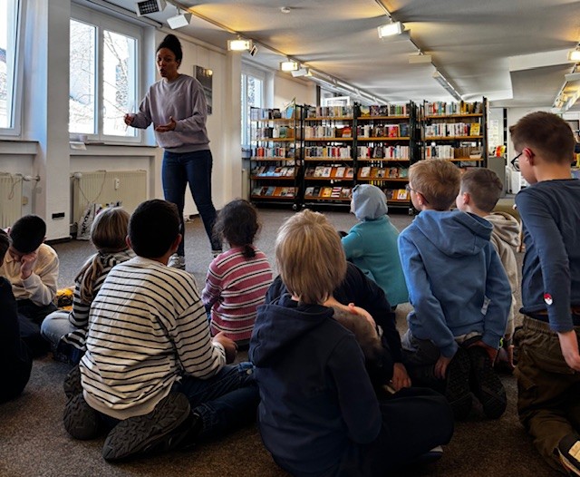 Eine Gruppe Kinder sitzt in einer Bibliothek auf dem Boden und lauscht einer Vorlesung