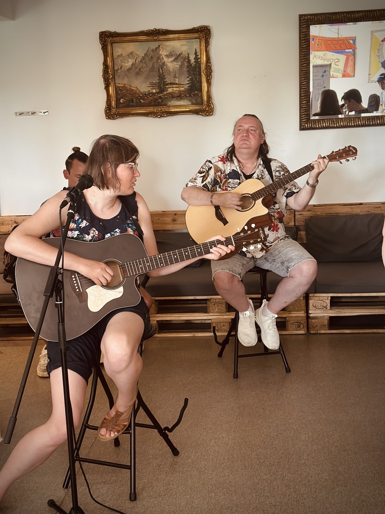 Links im Vordergrund sitzt eine junge Frau auf einem Stuhl und spielt Gitarre. Im Hintergrund sitzt ein Mann auf einer Bank und spielt auch Gitarre