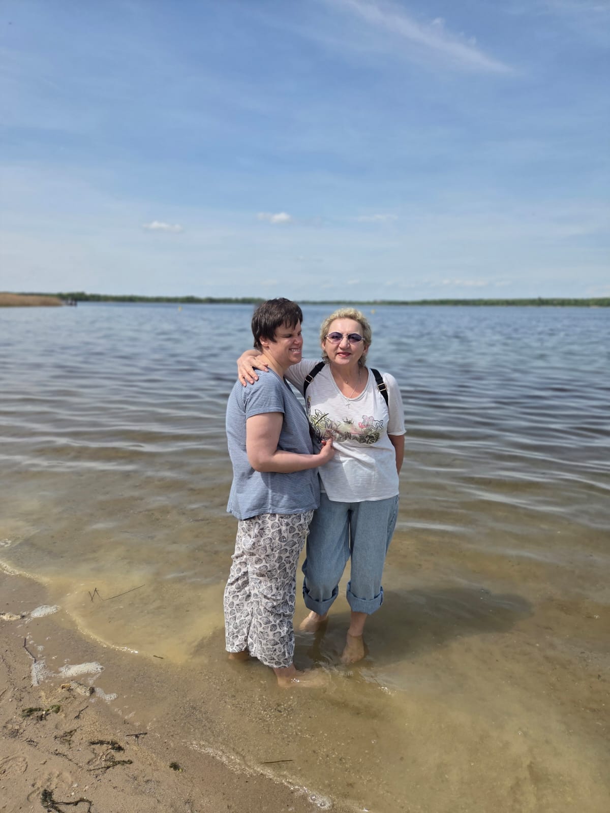 Zwei erwachsene Frauen stehen barfuß im seichten Wasser am Rand eines Sees. Die Frau links hat kurze, dunkle Haare und eine helle Haut. Sie trägt ein hellblaues T‑Shirt und eine weiße Hose mit dunkelgemustertem Druck. Ihr Körper ist leicht zur Frau rechts gedreht, und ihr rechter Arm liegt locker um deren Rücken.