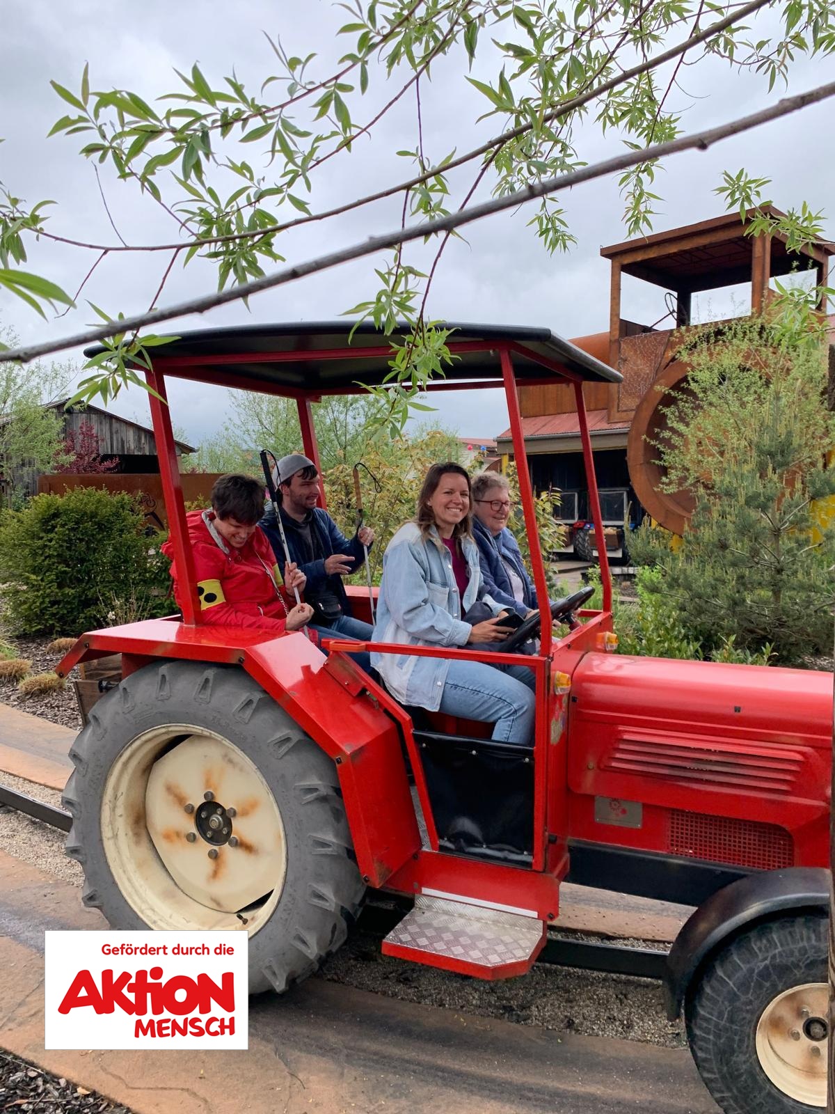 Roter Traktor mit überdachtem Anhänger auf einer Schiene in einer Parklandschaft. Auf dem Fahrersitz vorne rechts sitzt eine junge Frau mit mittellangen hellbraunen Haaren, hellerer Haut, hellblauer Jeansjacke und blauer Jeans; sie lächelt deutlich. Direkt hinter ihr sitzt eine weitere Person mit heller Haut, Brille, kurzem hellbraunen Haar und dunkelblauer Jacke, die nach vorne schaut. Links davon sitzen zwei weitere Personen nebeneinander: vorn eine Person mit heller Haut, dunklen kurzen Haaren, roter Jacke und gelbem Armband am linken Oberarm, die nach unten zu einem Gegenstand in den Händen blickt; dahinter eine Person mit heller Haut, dunkler Kappe, dunkelblauer Jacke und Bart, die seitlich lächelt.